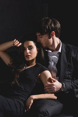 attractive brunette woman in black dress and handsome man in suit sitting on couch and hugging isolated on black