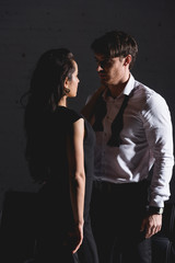 man in white shirt and woman in black dress standing close and looking to each other on dark background