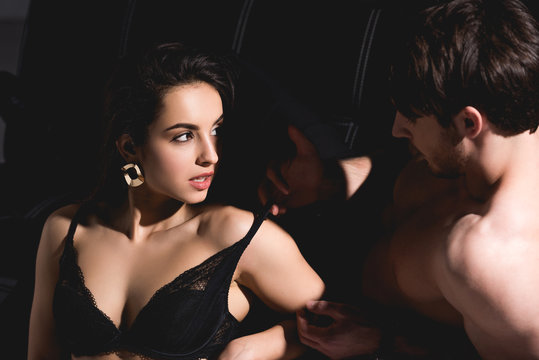 handsome man on couch undressing brunette woman in lingerie sitting on floor on black background