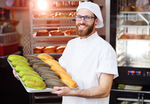 Young Cute Baker In White Uniform Holding A Tray With Colored Rolls For Hot Dog On The Background Of Bakery Or Bread Factory