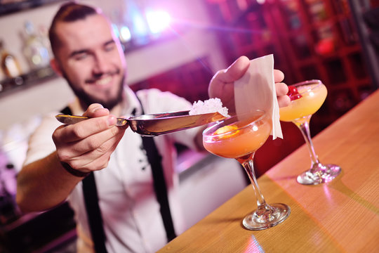 the bartender holding two glasses with an orange alcoholic cocktail smiles against the night club or bar. Bartender Show.