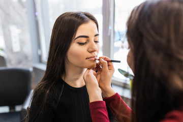 Makeup artist applies red lipstick . Beautiful woman face. Hand of make-up master, painting lips of young beauty model girl . Make up in process