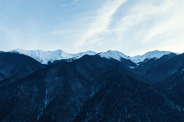 Aerial view of the mountain range against the blue sky. Panorama of mountains with snowy tops. Broad-leaved and pine forests in the winter evening sunset. Mountains in the winter. Ski resorts. Drone