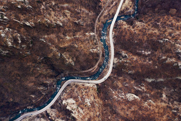 Aerial view of the mountain range, the flowing river and the paved highway. Winding road for cars among the mountains. Deciduous forest in the winter evening. Drone