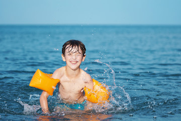Happy smiling boy bathes in the sea near the shore with splashes of water. Wet hair. Orange inflatable swim sleeves. A child at the sea, rest in the summer, swimming in the sea. Summer school holidays