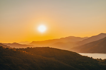 Panorama of the peaks of the beautiful tree-covered mountains at sunset on a sunny day in summer. Bright large orange sun disc. Solar reflection. A flash of the sun. Lens flare. Relax for tourist