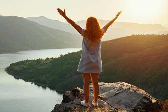 A relaxed red-haired woman in a dress is standing on top of a high mountain against the backdrop of a large mountain lake and spinning with happiness, raising her hands up. Bright sunset on the lake