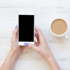 Female hands using smartphone and drinking latte over white wooden table, top view. Flat lay, from above.