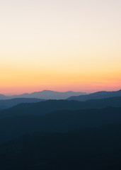 Panorama of the peaks of beautiful mountains covered with trees at sunset on a sunny day in summer. Orange glow on the sky after full sunset. Dark silhouettes of mountains. Solar reflection