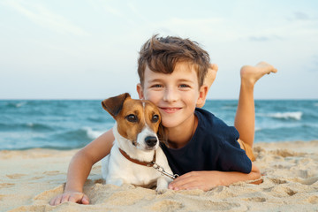 Happy boy hugging his dog breed Jack Russell terrier at the seashore against a blue sky close up at sunset. Best friends rest and have fun on vacation, play in the sand against the sea summer