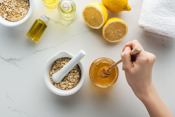 partial view of female hand with honey stick, pounder with oat flakes, lemons and different natural ingredients on white surface