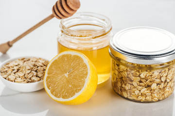 selective focus of glass jars with honey and oat flakes, lemon and honey stick on white surface