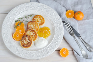 Cheesecakes with citrus jam and sour cream. Next to the plate is a cloth and cutlery. The composition is complemented by mandarin slices. Light wooden background. Close-up. View from above.