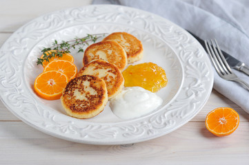 Cheesecakes with citrus jam and sour cream on a white plate. Next to the plate is a cloth napkin and cutlery. The composition is complemented by slices of mandarin. Light wooden background. Close-up.