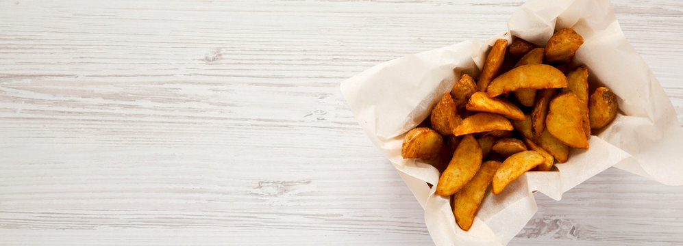 Fried potato wedges in paper box on a white wooden background. Overhead, top view. Copy space.