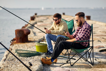 leisure and people concept - friends with smartphones fishing on pier at sea