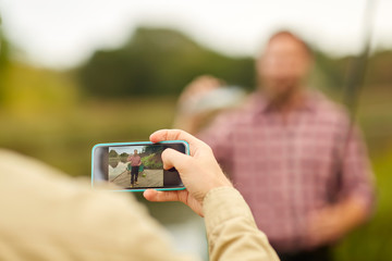 fishing, leisure and people concept - friend photographing fisherman by smartphone at lake
