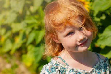 Close-up. Lifestyle photo. Cute cheerful redhead girl playing on the meadow, field. Beautiful little girl in the meadow near sunflowers and smiling. Soft sunlight.