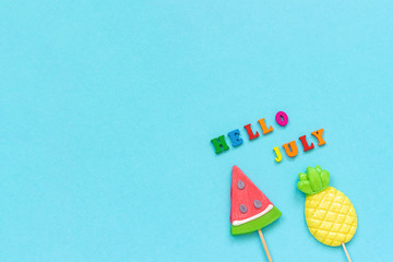 Hello July colorful text, pineapple and watermelon lollipops on stick on blue paper background. Concept vacation or holidays Creative Top view Copy space Template Greeting card, postcard