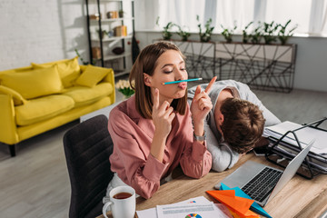 funny girlfriend sitting near boyfriend sleeping at desk near laptop and cup with drink