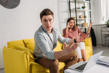 Naklejka premium selective focus of man in glasses gesturing near laptop with girlfriend talking on smartphone while sitting on sofa on background