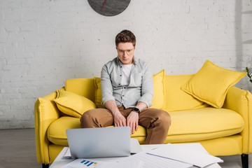 Fototapeta premium tired man in glasses sitting on sofa and looking at laptop at home