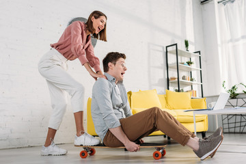 cheerful girlfriend pushing skateboard of boyfriend in glasses at home
