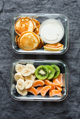 Healthy kids school, walk, picnic lunch box - pancakes with sour cream and banana, kiwi, tangerine fruit. Delicious snack on a grey background, top view