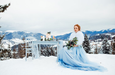 A comely girl in a blue dress with bouquet in hands sitting on a chair against a background of winter mountains