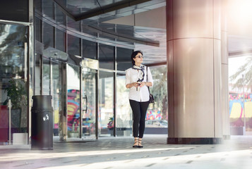 Fototapeta na wymiar Beautiful European caucasian woman coming out a city building wearing a white shirt and black tight pant and a scarf holding a coffee container and hand bag 