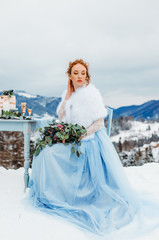 Fototapeta na wymiar Amazing red-haired girl with a bouquet in her hands sitting on a chair against the backdrop of winter mountains