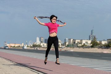 Fototapeta na wymiar Beautiful European caucasian woman jumping and laughing on the side of a city highway wearing pink shirt and black tight pant and a playing with her scarf 