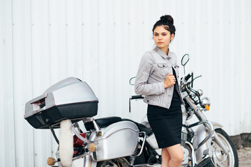 Young pretty cute brunette girl dressed in a little black dress and a jacket standing near a motorcycle
