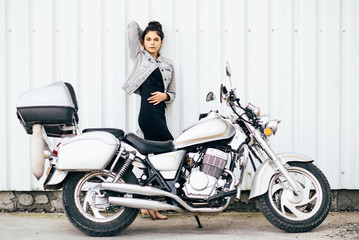 Fototapeta na wymiar Young pretty cute brunette girl dressed in a little black dress and a jacket standing near a motorcycle