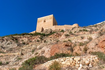 Fototapeta premium A steep rocky hill leading up to the square tower of Lorca Castle framed by an ultra blue sky in southern Spain