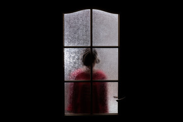 Dark silhouette of girl in red behind glass. Locked alone in room behind door on Halloween. Nightmare of child with aliens, monsters and ghosts. Evil in home. Inside haunted house. Supernatural.