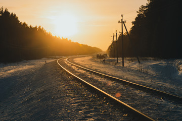 Fototapeta na wymiar Empty railroad in winter forest at sunset. Dark silhouettes of trees in backlight of dawn. Railway track in golden sunlight. Gold sunbeams. Rails on snow in sun rays. Wonderful atmospheric landscape.