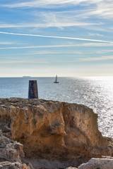 A location marker situated along the rocky coastline of southern Spain looks over the tranquil water of the Mediterranean where a small sailboat sails past and a much larger ship sits on top of the ho