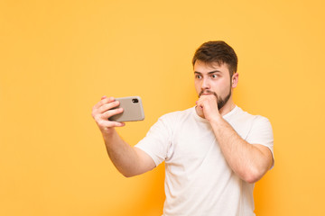 Shocked man dressed in a white T-shirt stands on a yellow background with a smartphone in his hands, looks at the smartphone screen, covered his mouth with amazement, isolated on a yellow background