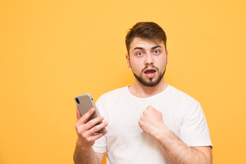 Emotional man with a beard, wearing a white T-shirt, stands on a yellow background with a smartphone in his hands and looks surprised at the camera. Isolated. Copyspace