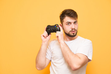 Handsome man with a beard wearing a white T-shirt,posing with a joystick in his hands on a yellow background. Portrait of an adult gamer with a gamepad in his hands, looking in camera. Gamer concept