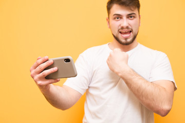 Joyful bearded man holds the smartphone in his hands and rejoices in victory, the focus on the smartphone.Adult man in a white T-shirt with a smartphone in his hands is isolated on a yellow background