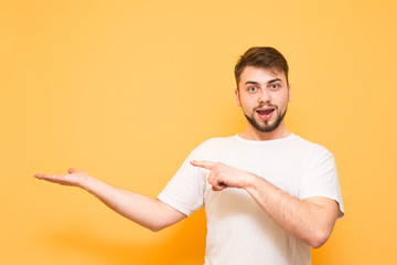 Emotional man in a white T-shirt stands on a yellow background, shows his hands in an empty place and looks surprised at the camera. Funny man with beard shows on copyspace