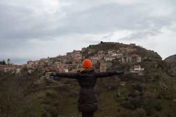 Fototapeta na wymiar Woman wearing an orange hat stands with wide open hands in front of the Greek village Vytina, during a cold, cloudy, grey winter day