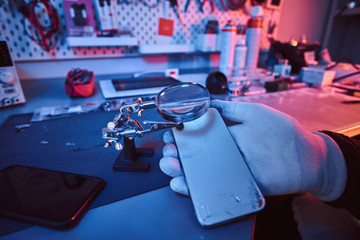 Electronic technician holds a modern smartphone with a broken body, carefully examines the damage using a magnifying glass sitting at the desk in the repair shop. Illumination with red and blue lights