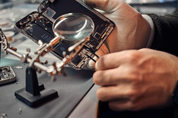 Repairman uses magnifier and tweezers to repair damaged smartphone. Close-up photo of a disassembled smartphone.