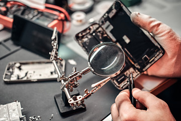 Repairman uses magnifier and tweezers to repair damaged smartphone. Close-up photo of a disassembled smartphone.