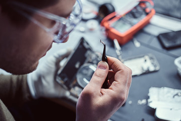 Electronic technician mending a broken phone, looking closely at the little bolt holding it with tweezers in the repair shop