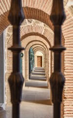 A series of bricked arches seen through a blurred cast iron fence leading to a wooden door on the far-side.