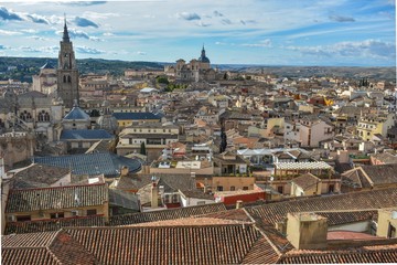 A tiled rooftop view of Toledo, Spain with the towers of the cathedral and monastery peaking out from amongst the closely spaced buildings. 